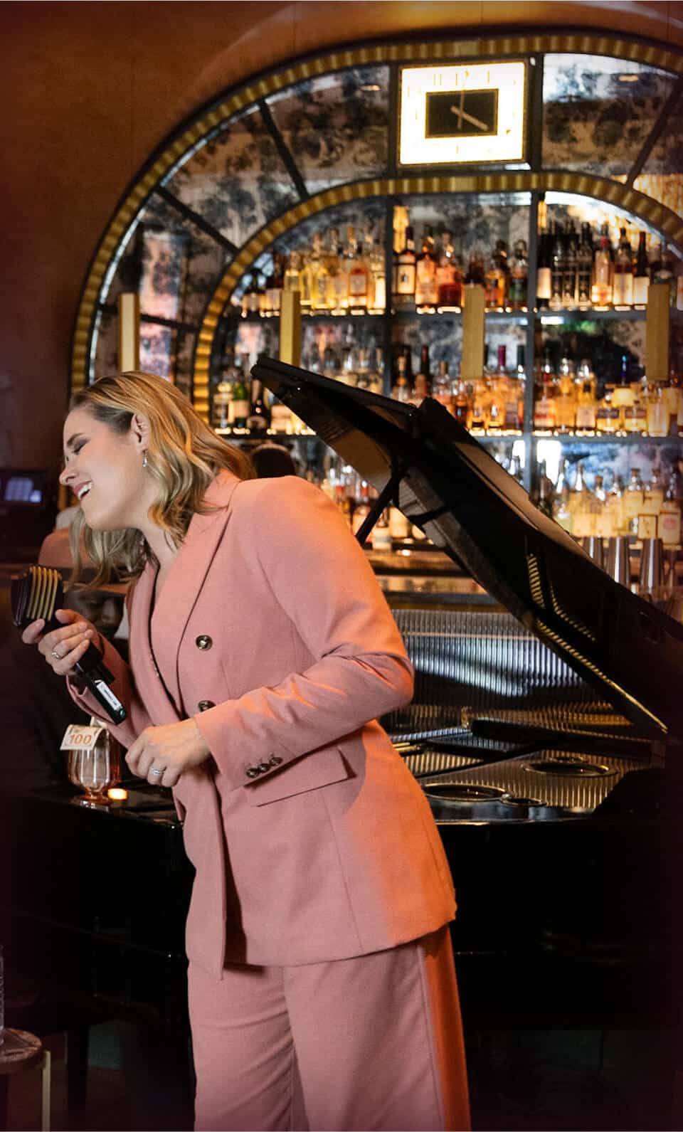Elegant woman in pink suit enjoying jazz performance at a sophisticated bar with grand piano, jazz music, and stylish ambiance, promoting luxury nightlife experience.