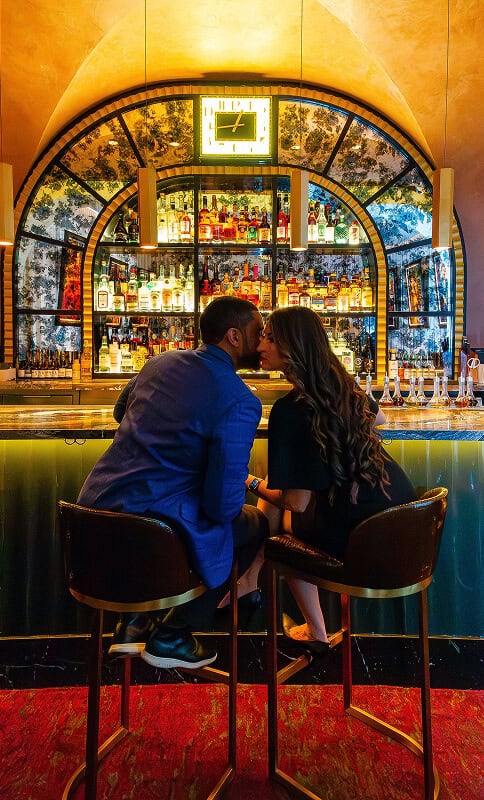 Vibrant bar scene with a couple kissing at the counter, illuminated by colorful bottles and warm lighting, showcasing nightlife and romantic moments in an upscale lounge setting.