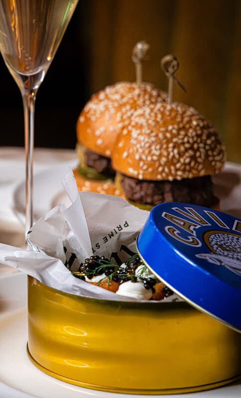 Order of The Ace gourmet burger with sesame seed bun, fresh toppings, and side snacks served in a stylish tin container with a drink, representing premium dining experience.