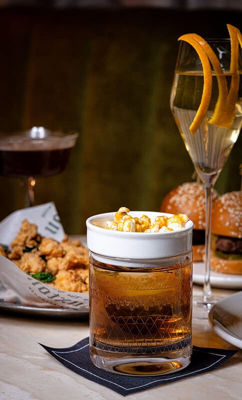 Golden whiskey with caramel popcorn in a glass tumbler, surrounded by gourmet burgers, a champagne flute with lemon, and fried chicken, creating an elegant bar-food setting for the 